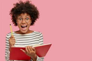 Overjoyed black woman holds textbook and pencil, exclaims with amazement, creats bestseller, wears round glasses, poses against pink wall with copy space on right side. Well, I get good idea for poem